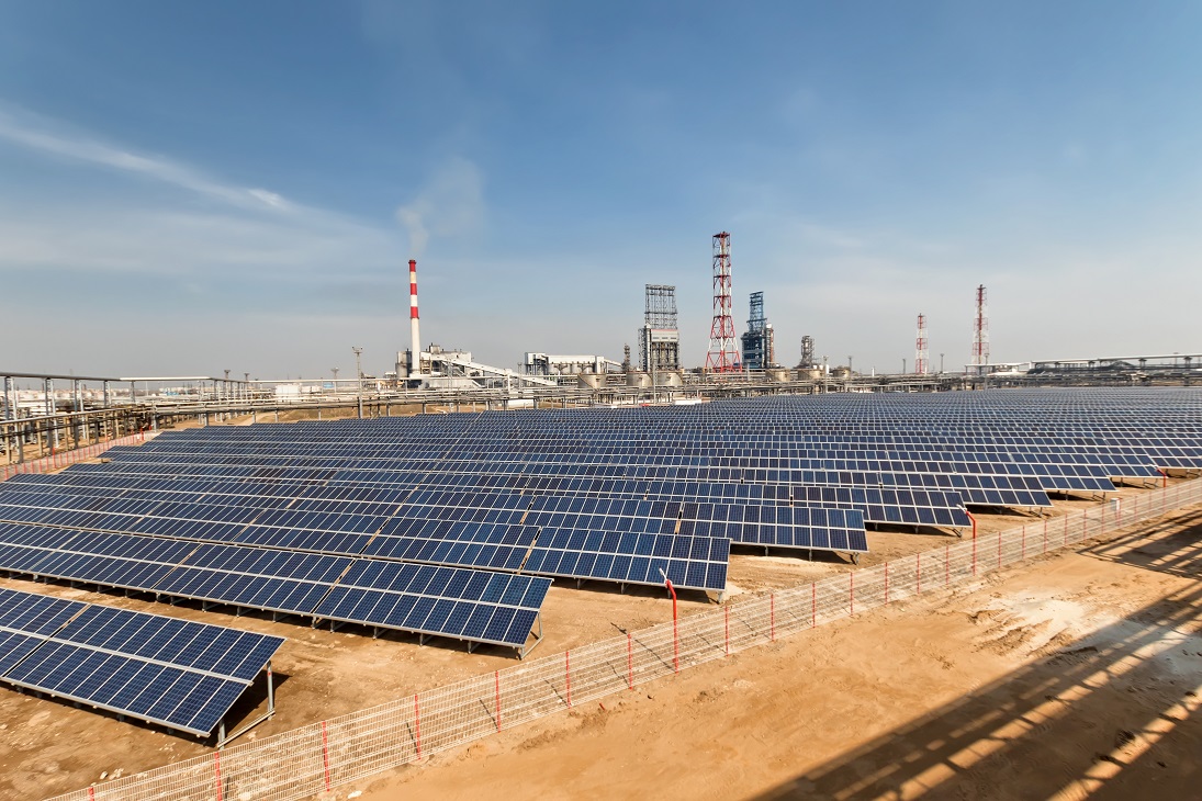 Ground-mounted solar PV array beside an industrial facility under a clear sky